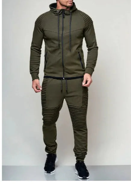 A man is dressed in an olive green tracksuit with a modern, slim-fit design. The hoodie features ribbed detailing on the shoulders, a front zipper, and black drawstrings. The matching joggers have ribbed accents on the thighs and knees, along with black drawstrings at the waist. He is wearing black sneakers with white soles. The neutral background highlights the stylish and sporty appearance of the outfit.