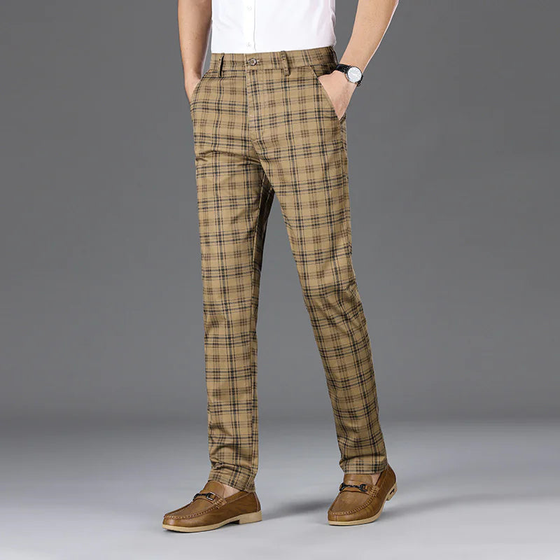 A man wearing slim-fit, beige plaid trousers with a black checkered pattern. The trousers have a classic button and zipper closure, belt loops, and side pockets. He pairs them with a white button-up shirt tucked in, brown leather loafers with a decorative metal accent, and a black wristwatch. The background is a simple gray backdrop, emphasizing the stylish and sophisticated outfit.