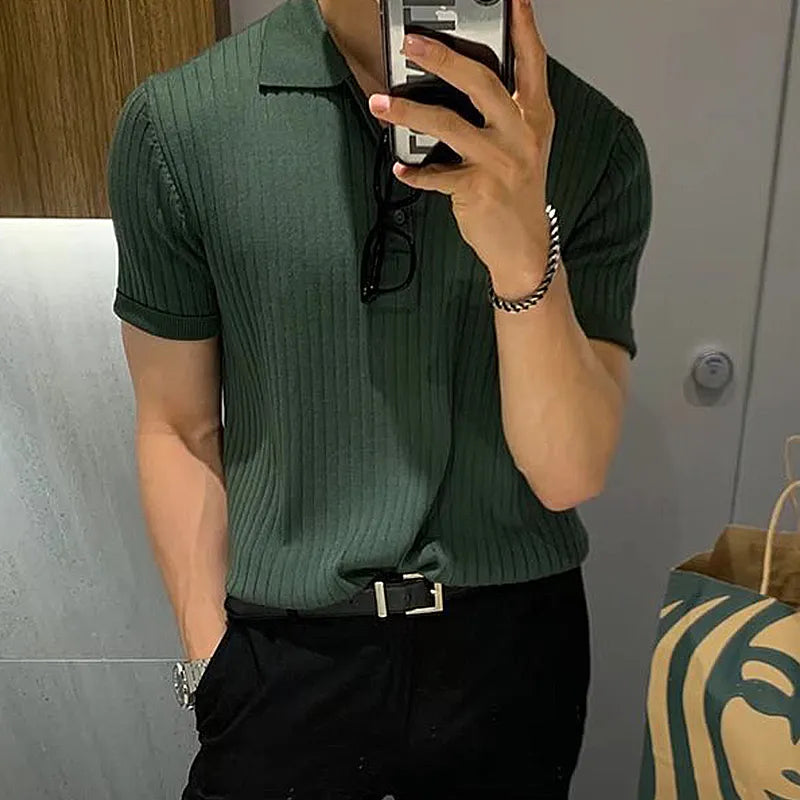 A stylish man takes a mirror selfie in a modern restroom, partially covering his face with his smartphone. He is wearing a fitted green ribbed polo shirt tucked into black trousers with a black belt. A pair of glasses hangs from the collar of his shirt. He accessorizes with a silver watch on his left wrist and a silver bracelet on his right wrist. The background features a wooden panel and a marble wall, with a paper shopping bag visible on the right side of the frame. The lighting is warm and soft...