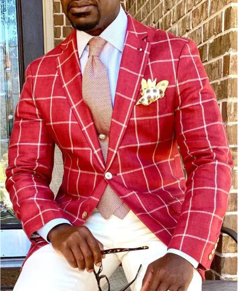 A stylish man sits outdoors against a brick wall, dressed in a red windowpane blazer with white grid patterns. He pairs it with a crisp white dress shirt, a textured salmon-colored tie, and a matching waistcoat. His pocket square, adorned with decorative elements, adds a unique touch to his sophisticated ensemble. He completes the look with white trousers. In his hand, he holds a pair of glasses, exuding confidence and elegance. The natural lighting enhances the rich tones of his outfit.