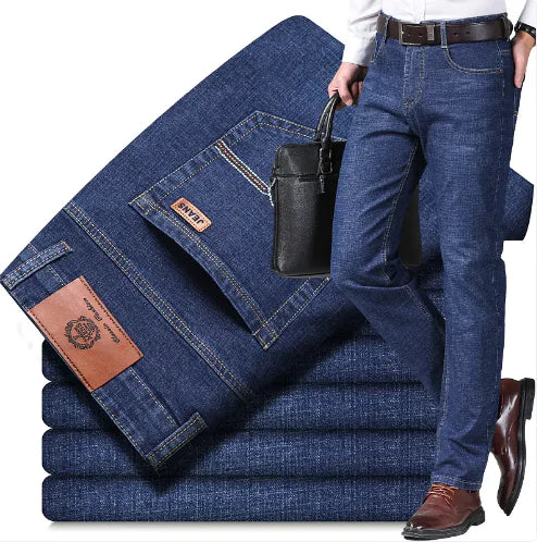 A man dressed in formal attire showcases a pair of classic straight-cut blue jeans. He pairs the jeans with a white dress shirt, brown leather belt, and matching brown dress shoes. He is holding a black leather briefcase, suggesting a smart-casual or business-casual style. Behind him, several neatly folded pairs of the same denim jeans are displayed, highlighting the fabric quality and design details, including leather brand patches and stitching.