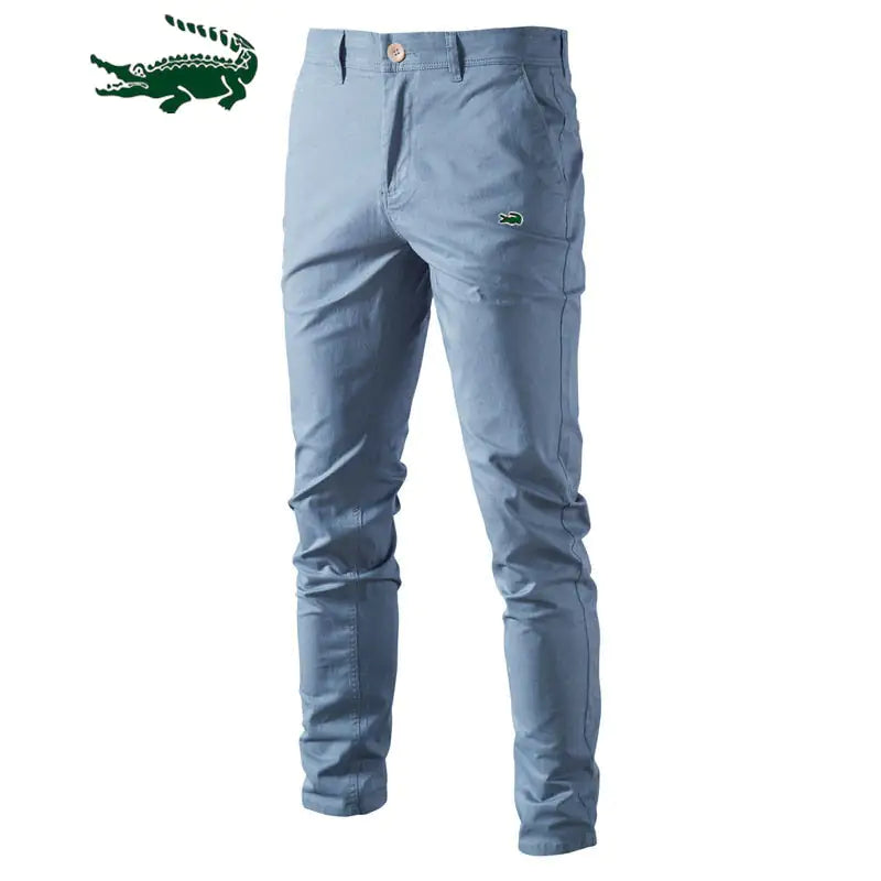 A pair of slim-fit khaki pants with a button and zipper closure, featuring a small green crocodile logo on the upper left thigh. The pants have belt loops and side pockets, with a slightly wrinkled appearance. A larger green crocodile logo is displayed in the top left corner of the image.