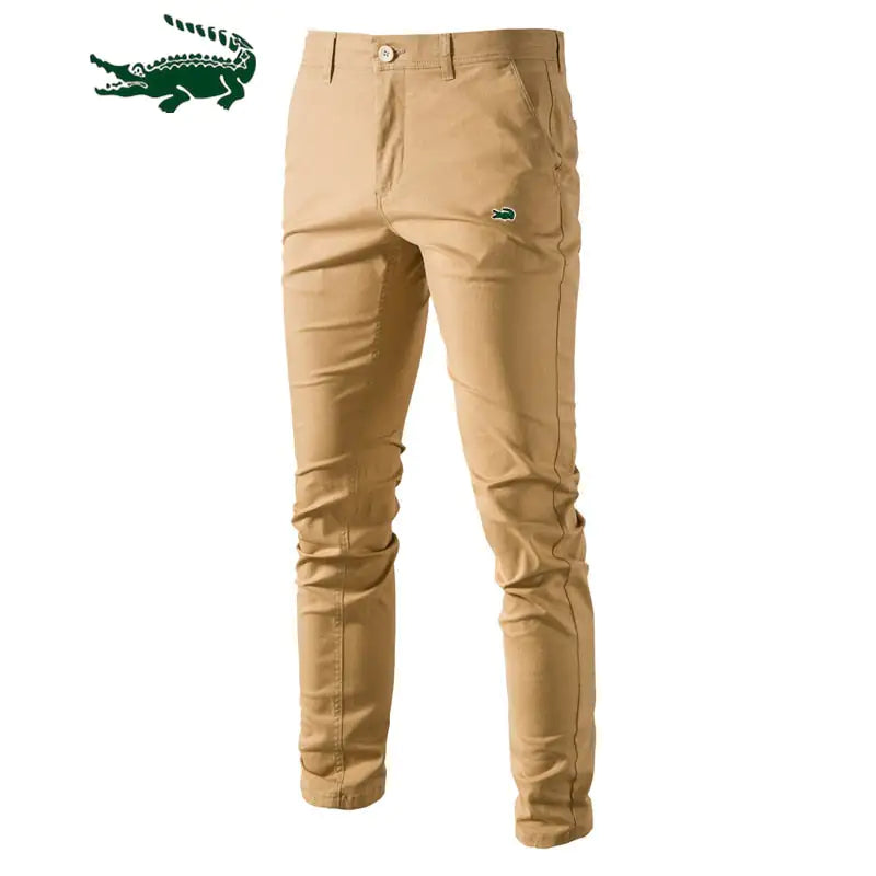 A pair of slim-fit khaki pants with a button and zipper closure, featuring a small green crocodile logo on the upper left thigh. The pants have belt loops and side pockets, with a slightly wrinkled appearance. A larger green crocodile logo is displayed in the top left corner of the image.