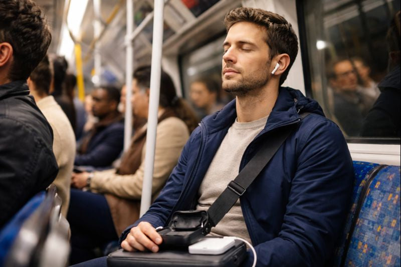 A man on a packed commuter train in London uses noise-cancelling earbuds, eyes closed, while a USB-C charging case sits on his lap next to a power bank.