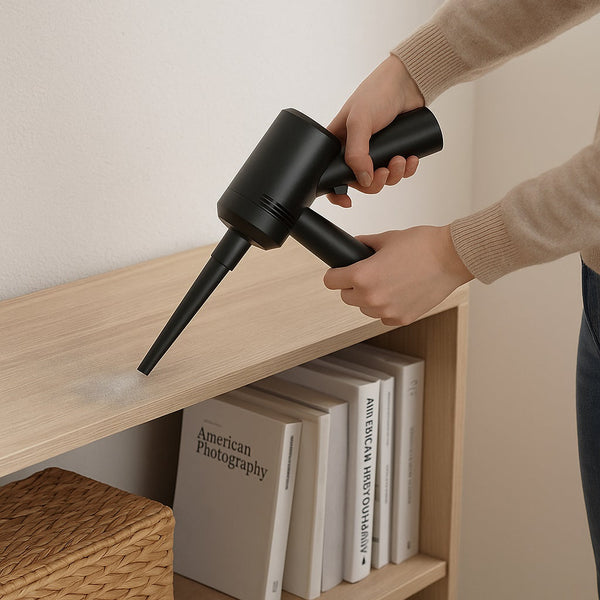 A person using a compact duster blower vacuum cleaner to clean dust from a white shelf in a modern, well-lit living room.