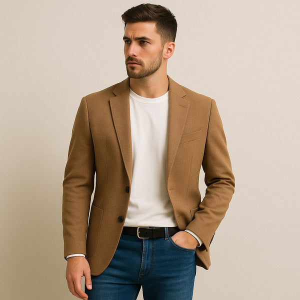 A young Caucasian male model wearing a camel blazer over a white T-shirt and blue jeans, posing confidently against a neutral off-white background with a clean, modern look.