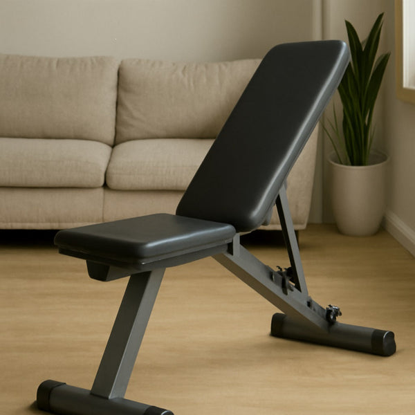 A black adjustable weight bench with a cushioned backrest and metal frame in a bright living room. Behind it is a beige sofa and a green snake plant, with soft natural light coming through a window, creating a peaceful and inviting space.