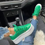 A pair of vibrant green platform sneakers worn by a person sitting in a car, with ripped blue jeans and a gold ankle bracelet. The sneakers feature white soles and are placed on the car’s floor.