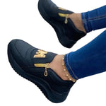 A pair of black slip-on casual shoes with gold zipper accents and decorative gold embellishments across the top. The shoes are shown worn with blue skinny jeans and a delicate ankle bracelet, featuring thick black soles and a sleek low-cut design.