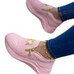 Person wearing pink slip-on sneakers with a chunky sole, a gold crown emblem on the upper, and a small gold zipper detail on the side, paired with blue jeans and an ankle bracelet.