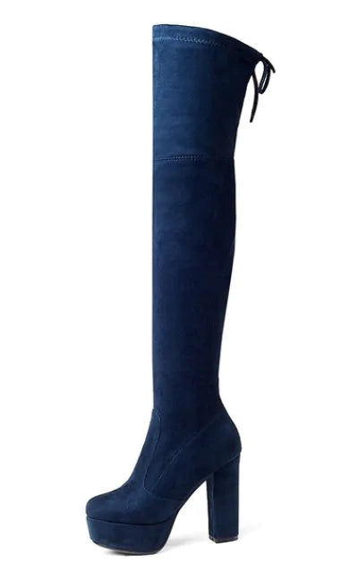 A woman wearing black high-heeled, over-the-knee boots with an open lattice cutout design running up the back, revealing skin between the horizontal straps. She stands indoors on a light wooden floor near a white ornate console table.