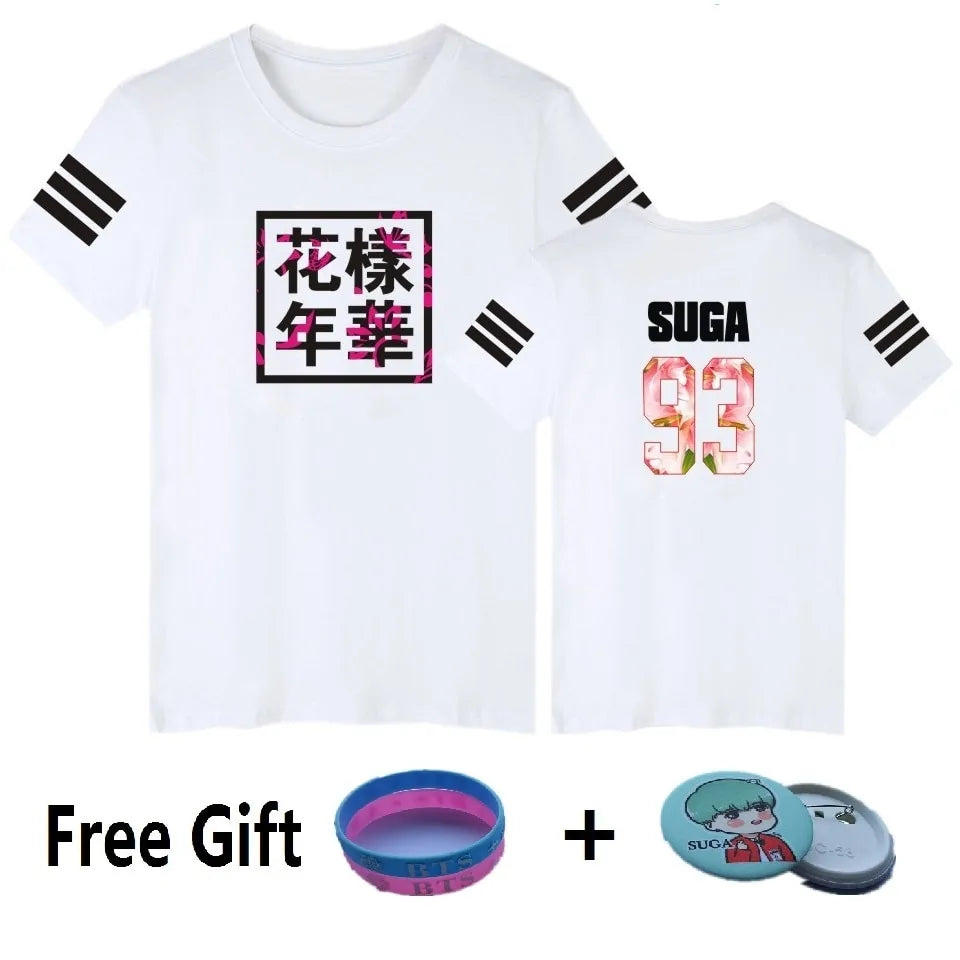 A white short-sleeve graphic T-shirt shown from both the front and back. The front features a square black-bordered design with bold black Asian characters over a pink floral pattern, while the back displays the name “SUGA” in black above the number “93” in a pink floral print. Both sleeves have three black stripe accents. At the bottom, the image also shows free gift items: a blue-and-pink wristband and a round character pin badge.