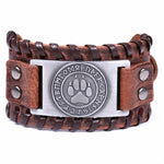 Brown braided leather cuff bracelet with metal studs and a central silver plate featuring a paw print emblem surrounded by rune-like symbols, shown on a white background.
