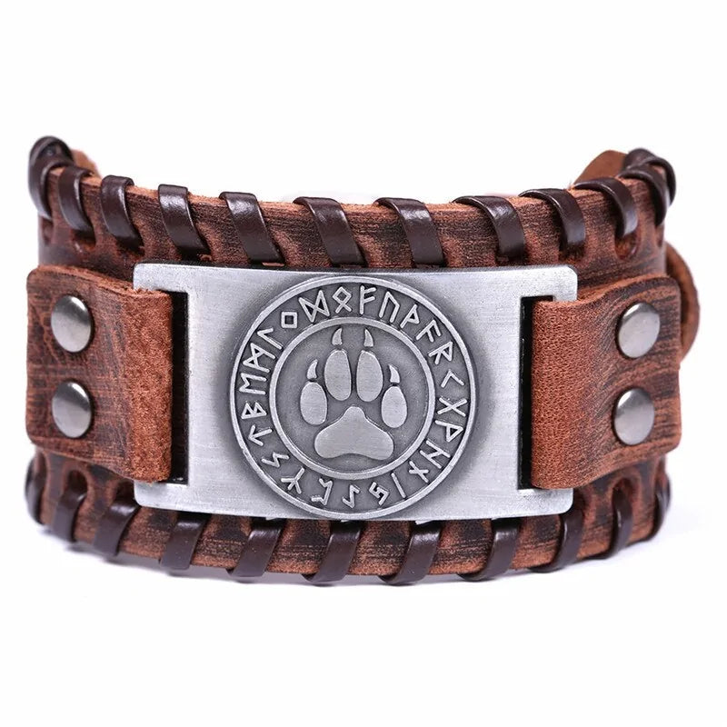 Brown braided leather cuff bracelet with metal studs and a central silver plate featuring a paw print emblem surrounded by rune-like symbols, shown on a white background.