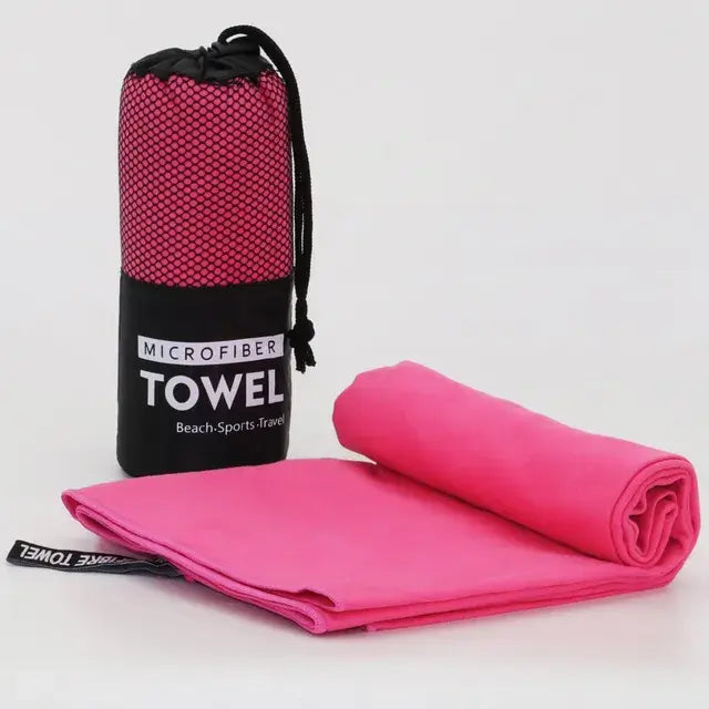 Bright pink microfiber towel partly rolled and folded on a white surface, next to a matching pink towel stored in a black-and-pink mesh drawstring pouch labeled “MICROFIBER TOWEL Beach·Sports·Travel.”