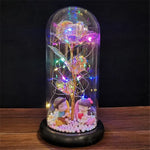 Red rose displayed under a clear glass dome on a wooden base, wrapped with warm white fairy lights and glowing on a rustic wooden surface against a dark background.