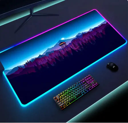 RGB LED gaming mouse pad on a desk, glowing around the edges and printed with a landscape design, shown with a backlit keyboard and mouse.