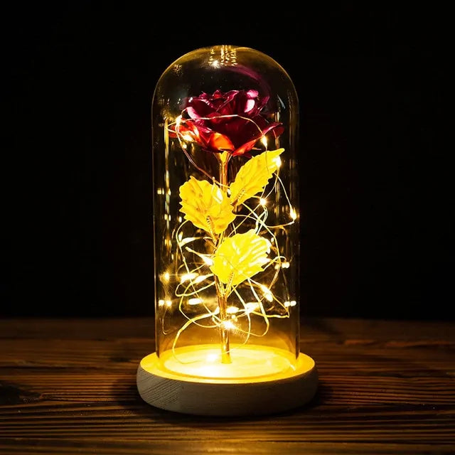 Glowing artificial red rose displayed inside a clear glass dome, wrapped with warm white fairy lights and set on a round wooden base against a dark background.