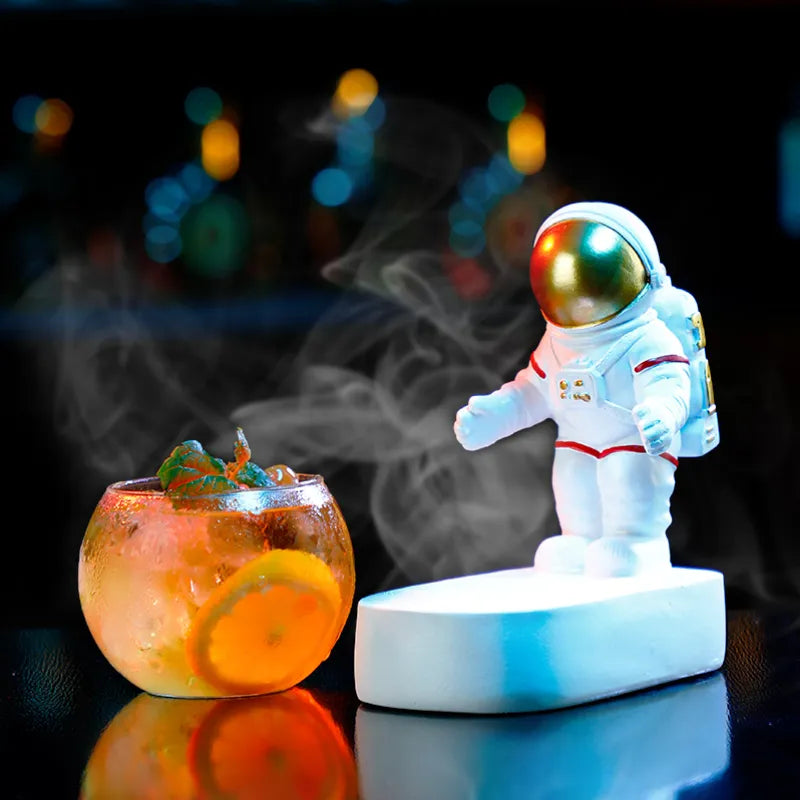 Small astronaut figurine on a white base beside a glass of iced citrus drink, with misty vapor swirling in front of a dark, blurred background with colorful lights.