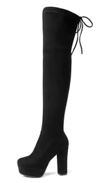 A woman wearing black high-heeled, over-the-knee boots with an open lattice cutout design running up the back, revealing skin between the horizontal straps. She stands indoors on a light wooden floor near a white ornate console table.