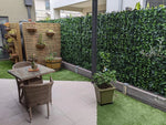 Small outdoor patio with a glass-top table and wicker chairs on tiled flooring, bordered by artificial grass, a wooden slat planter wall with hanging baskets, and a dense green ivy privacy fence.