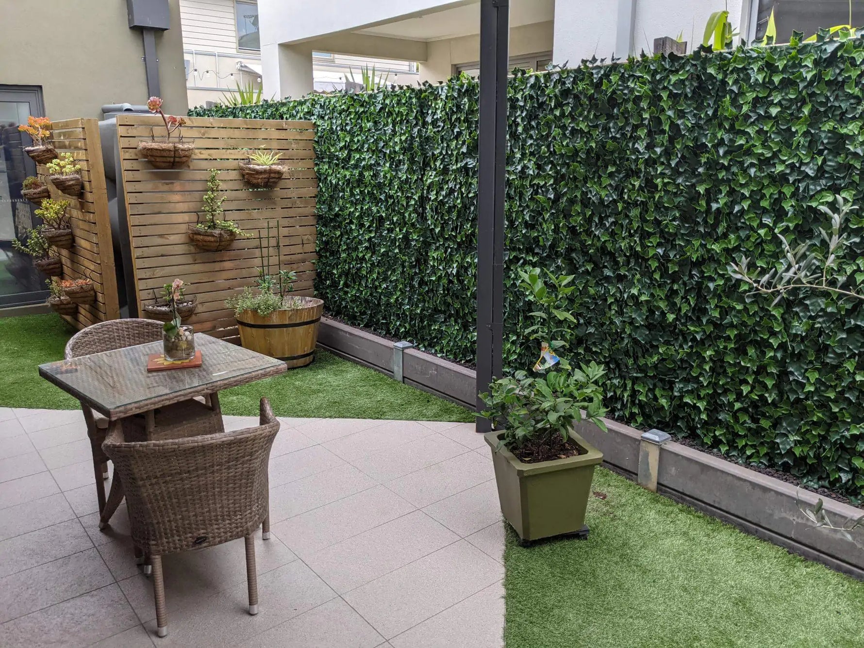 Small outdoor patio with a glass-top table and wicker chairs on tiled flooring, bordered by artificial grass, a wooden slat planter wall with hanging baskets, and a dense green ivy privacy fence.