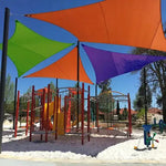 A colorful playground with climbing structures and slides is shaded by large triangular fabric canopies in orange, green, blue, and purple, set over white sand under a clear blue sky.