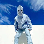 Person in a light blue hoodie with a skull-print zipped hood, posing against a bright blue sky.