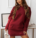 Woman wearing a long-sleeve, oversized maroon hoodie dress with a front kangaroo pocket and gold drawstrings. She is holding a light-colored handbag and posing indoors with a soft smile. The outfit has a casual, cozy look perfect for lounging or casual outings.