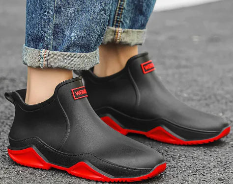 A pair of black waterproof ankle rain boots worn with cuffed jeans. The boots have a sleek, modern design with thick, textured soles and a branded label on the front that reads "WEMAM." The setting is an outdoor pavement, suggesting the boots are suited for wet or rainy weather.