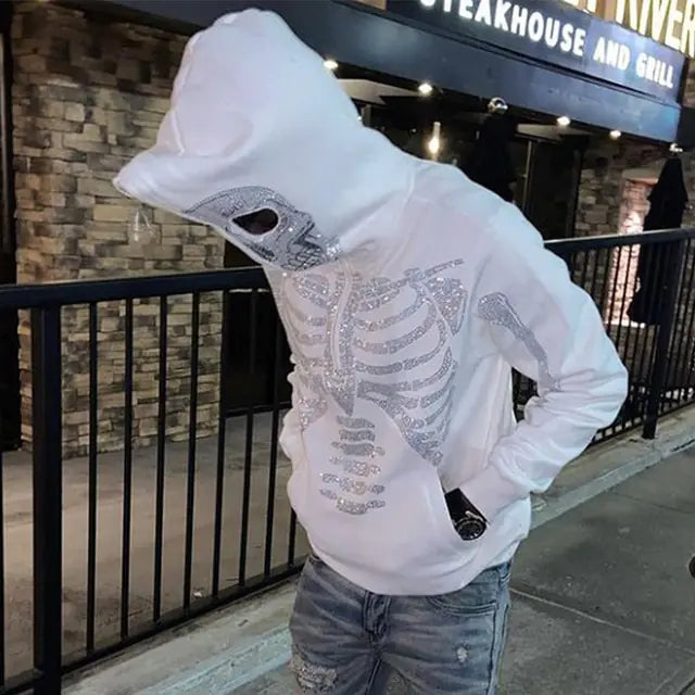 White zip-up hoodie laid flat on a concrete surface, featuring bold white skeleton graphics on the chest and arms, plus skull designs on the hood.