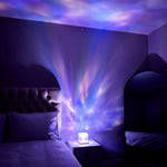 Dim bedroom lit by a small cube lamp on a nightstand, projecting shifting blue and purple galaxy-like patterns across the wall and ceiling beside a bed with pillows.