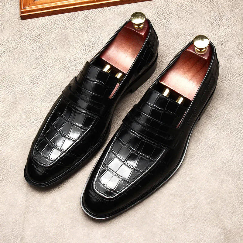 A pair of elegant burgundy leather loafers with a glossy finish. The shoes feature gold-toned metal horsebit detailing across the uppers and are displayed with wooden shoe trees inserted. They have a sleek, elongated silhouette with subtle stitching along the toe cap. The loafers are placed on a dark surface, partially framed by a wooden picture frame, emphasizing their classic and sophisticated style.