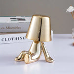 Small gold-colored table lamp shaped like a crawling figure holding a lampshade, sitting on a desk with stacked books and a small vase in the background.