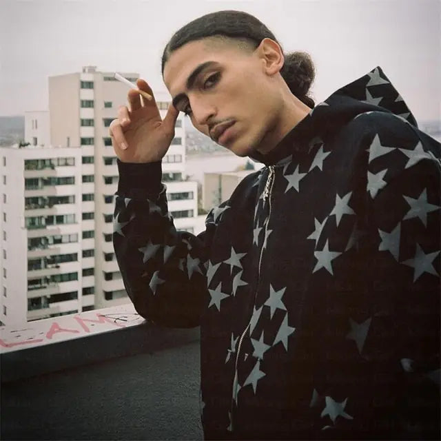 Person on a rooftop wearing a black hoodie with an all-over star pattern, holding a cigarette near their face with city buildings in the background.