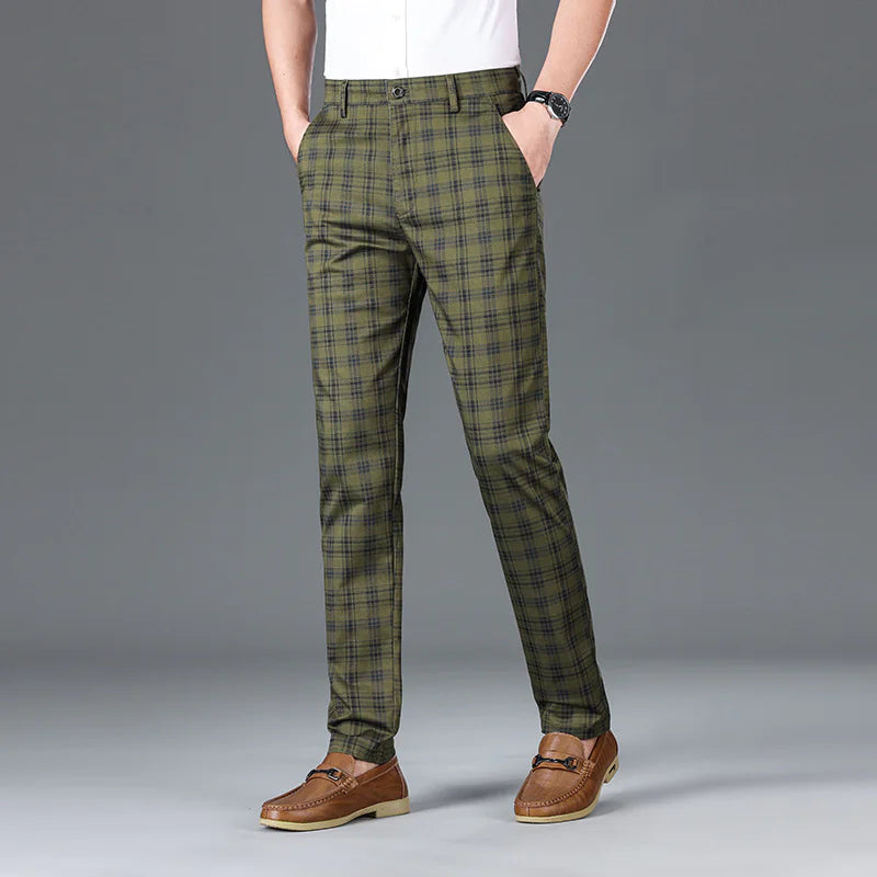 A man wearing slim-fit, beige plaid trousers with a black checkered pattern. The trousers have a classic button and zipper closure, belt loops, and side pockets. He pairs them with a white button-up shirt tucked in, brown leather loafers with a decorative metal accent, and a black wristwatch. The background is a simple gray backdrop, emphasizing the stylish and sophisticated outfit