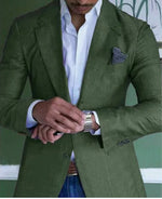A well-dressed man adjusts his silver wristwatch while wearing a tailored olive green blazer over a crisp white dress shirt. A dark gray pocket square is neatly tucked into the blazer’s breast pocket. He pairs the outfit with blue jeans and a brown leather belt, blending formal and casual styles. The background features a green wall with white trim and subtle decorative elements, complementing the blazer's earthy tone.