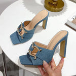 Pair of denim-blue square-toe mule heels with high block heels and gold horsebit-style hardware across the straps, shown on a white tabletop with one shoe held in a hand.