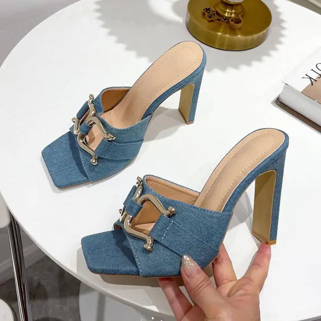 Pair of denim-blue square-toe mule heels with high block heels and gold horsebit-style hardware across the straps, shown on a white tabletop with one shoe held in a hand.