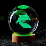 The image shows a glowing 3D-etched model of an axolotl inside a crystal ball, illuminated in green with a wooden base. The intricate details of the axolotl's shape and features are highlighted, creating a stunning visual effect.