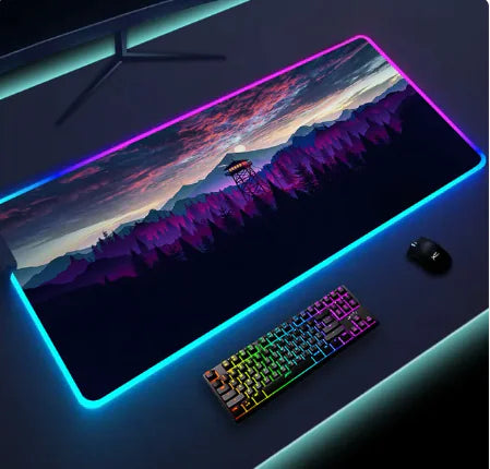 A dark gaming desk setup with a large RGB-lit mouse pad showing a grayscale mountain-and-forest scene, a rainbow-backlit keyboard, and a black mouse.