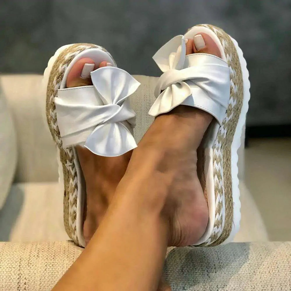 Person wearing white slide sandals with large bow details and braided espadrille-style platform soles, shown with feet resting on a sofa.