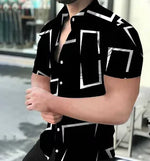 A muscular man wearing a stylish short-sleeved button-up shirt. The upper part of the shirt is purple with a geometric pattern resembling a honeycomb, while the lower part is solid black. The shirt is partially unbuttoned, revealing part of his chest. The background appears to be an outdoor urban setting with blurred reflections in a glass window and some chairs visible.