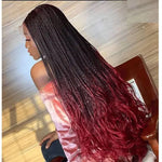 A person with long braided hair that transitions from black at the roots to a deep red at the ends, styled in loose waves. They are wearing a light pink shirt, and the photo is taken from the back, showing the full length of the hair.