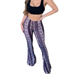 Woman wearing a black crop top and high-waisted, flared bohemian print pants with a mix of geometric and floral patterns in shades of purple, white, and maroon, posing with one hand on her hip