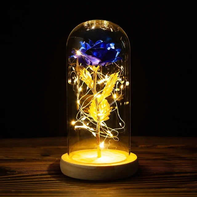 Blue rose with gold stem and leaves inside a glass dome, wrapped with warm white fairy lights, sitting on a wooden base against a dark background.
