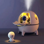 A white astronaut capsule-shaped air humidifier with a small figurine of an astronaut beside it, illuminated from within, emitting a mist, against a dark background.