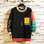 A stylish, oversized sweater with a black base color, featuring beige ribbed cuffs, collar, and hem. The right sleeve has a color-blocked design with red and green sections, while the left sleeve is black with "SUPNARIO" printed in bold beige letters. A yellow patch with an embroidered design and the words "YOUNG FOR" is sewn on the left chest. The sweater is displayed on a hanger against a textured stone wall background, with a long pendant necklace draped over it.