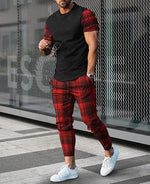 A stylish man is walking outdoors wearing a black t-shirt with red plaid sleeves, paired with matching red plaid jogger pants. He completes the look with white sneakers, a wristwatch, and sunglasses in hand. The background features a modern metal mesh wall and tiled pavement.
