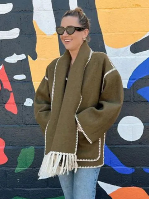 Dark wool coat with white blanket stitch trim, featuring front buttons, large patch pockets, and an attached matching scarf with fringe detailing draped over the shoulder. The coat is displayed against a plain white background.